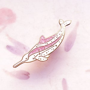 Op de afbeelding: Een witte en roze dolfijn emaille pin met goudkleurige accenten. De dolfijn heeft een roze glitterontwerp op zijn rug en staart, met kleine sterdetails. De pin is geplaatst tegen een zachtroze achtergrond.
