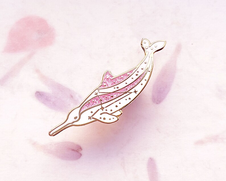 Op de afbeelding: Een witte en roze dolfijn emaille pin met goudkleurige accenten. De dolfijn heeft een roze glitterontwerp op zijn rug en staart, met kleine sterdetails. De pin is geplaatst tegen een zachtroze achtergrond.
