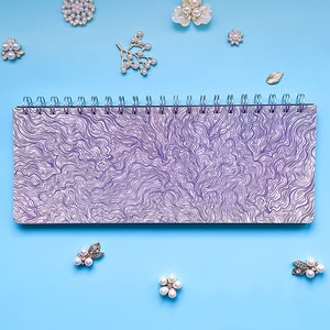 May include: A spiral bound notebook with a purple and white swirl pattern on the cover. The notebook is surrounded by small pearl and silver embellishments on a light blue background.