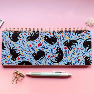 May include: A spiral-bound notebook with a blue cover featuring a pattern of black cats, red and yellow flowers, and green leaves. A gold binder clip and a light blue pen are on a pink surface.