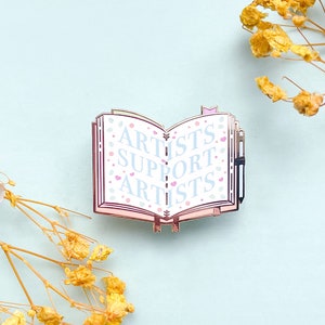 May include: A silver enamel pin shaped like an open book with the words "Artists Support Artists" in blue lettering on a white background. The book has pink edges and a small silver pen sticking out of the top.
