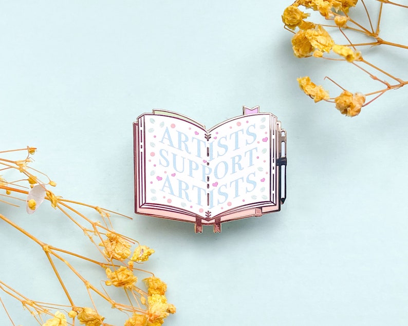 May include: A silver enamel pin shaped like an open book with the words "Artists Support Artists" in blue lettering on a white background. The book has pink edges and a small silver pen sticking out of the top.