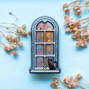 May include: A decorative window-shaped compact with a black cat silhouette on the sill. The window features a colourful, glittery design with shades of gold, orange, and blue. The compact is set against a light blue background with dried flowers.