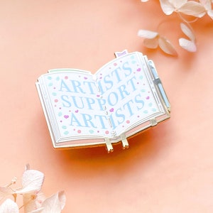May include: Gold-tone enamel pin shaped like an open book with the words "Artists Support Artists" in blue lettering on a white background. The book has a gold-tone bookmark and a small gold-tone heart on the front cover.