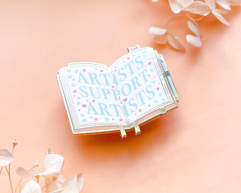 May include: Gold-tone enamel pin shaped like an open book with the words "Artists Support Artists" in blue lettering on a white background. The book has a gold-tone bookmark and a small gold-tone heart on the front cover.