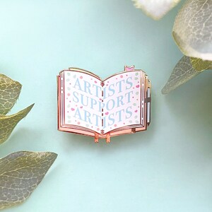 May include: A gold-toned enamel pin shaped like an open book with the words "Artists Support Artists" printed in blue on a pink background. The book has a pink and white floral pattern.