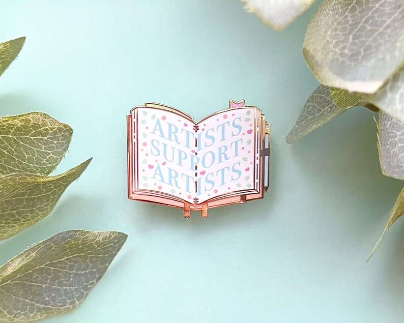 May include: A gold-toned enamel pin shaped like an open book with the words "Artists Support Artists" printed in blue on a pink background. The book has a pink and white floral pattern.