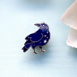 May include: A dark blue enamel pin in the shape of a crow, with silver-colored outlines and star details. The crow pin is set against a light blue background, with a white flower in the upper right corner.