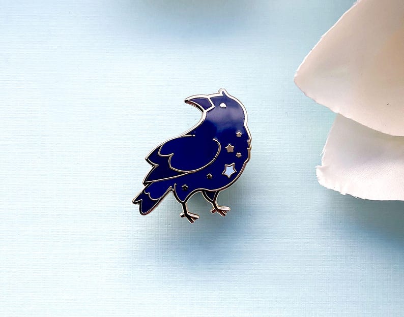 May include: A dark blue enamel pin in the shape of a crow, with silver-colored outlines and star details. The crow pin is set against a light blue background, with a white flower in the upper right corner.