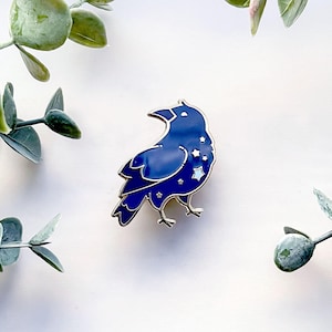 May include: A blue raven enamel pin with a silver outline and star details. The pin is set against a white background with green foliage.