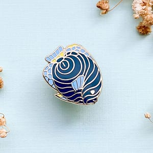 May include: A detailed enamel pin featuring a stylized blue and navy fish design with gold-colored trim. The fish has a unique pattern of curved lines and geometric shapes. The pin is set against a light blue background with dried flowers.