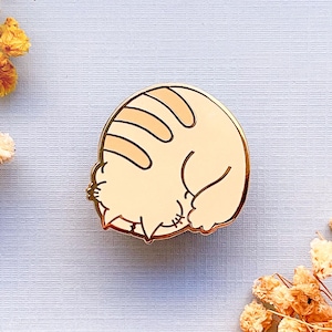 May include: Gold-tone enamel pin featuring a cartoon cat curled up in a ball, sleeping. The cat is light orange with white stripes and a pink nose.