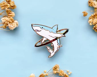 May include: A metal enamel pin featuring a white and light blue shark swimming through a dark metallic ring. The shark design includes small star and moon motifs on its body, with gold-toned metal outlines defining the shape of the shark and the ring.