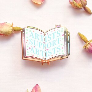 May include: Gold-toned enamel pin shaped like an open book with a blue and white background. The book has a pink ribbon bookmark and the words "ARTISTS SUPPORT ARTISTS" are written in blue on the pages.