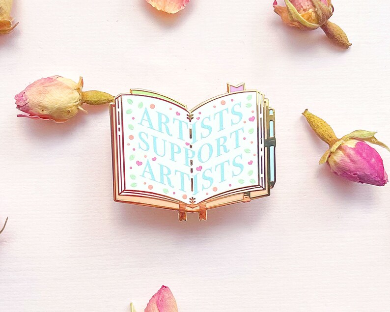 May include: Gold-toned enamel pin shaped like an open book with a blue and white background. The book has a pink ribbon bookmark and the words "ARTISTS SUPPORT ARTISTS" are written in blue on the pages.