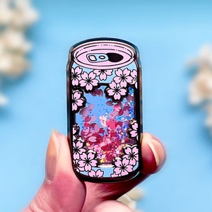 May include: A pink and black enamel pin shaped like a can of soda. The can is filled with pink and blue glitter and has a design of pink cherry blossoms around the top and bottom.