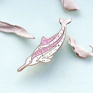 Op de afbeelding: Een witte en roze dolfijn emaille pin met gouden details. De pin heeft een glitterende roze streep en ster details. De dolfijn staat tegen een lichtblauwe achtergrond met gedroogde bladeren.