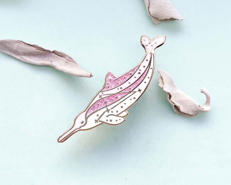 Op de afbeelding: Een witte en roze dolfijn emaille pin met gouden details. De pin heeft een glitterende roze streep en ster details. De dolfijn staat tegen een lichtblauwe achtergrond met gedroogde bladeren.