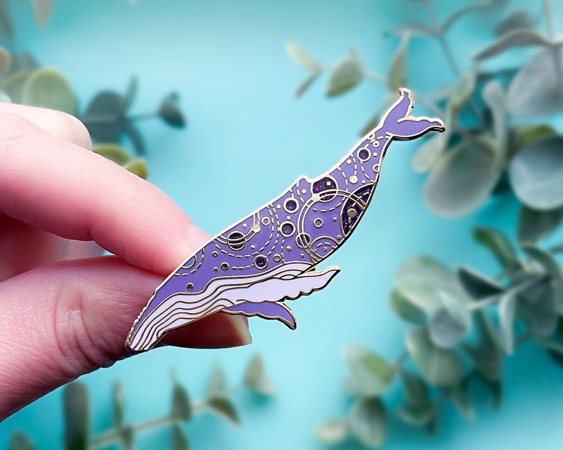 May include: Purple and gold enamel pin of a whale with a galaxy pattern. The whale is in a swimming position with its tail raised.