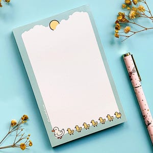 May include: A light blue notepad with a cloud and sun design at the top, and a row of yellow ducklings at the bottom. A pink pen with a mermaid design and gold accents is next to the notepad. Dried yellow flowers are in the background.