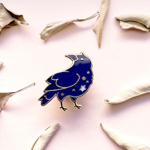 May include: A dark blue raven enamel pin with gold trim and star accents. The pin is set against a light pink background with dried leaves. The raven is facing left with its beak open.