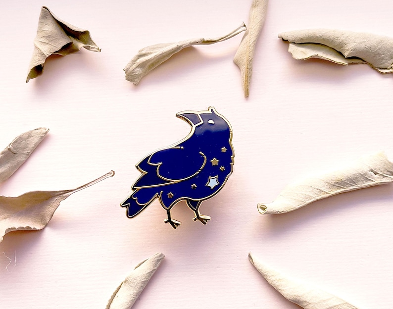 May include: A dark blue raven enamel pin with gold trim and star accents. The pin is set against a light pink background with dried leaves. The raven is facing left with its beak open.