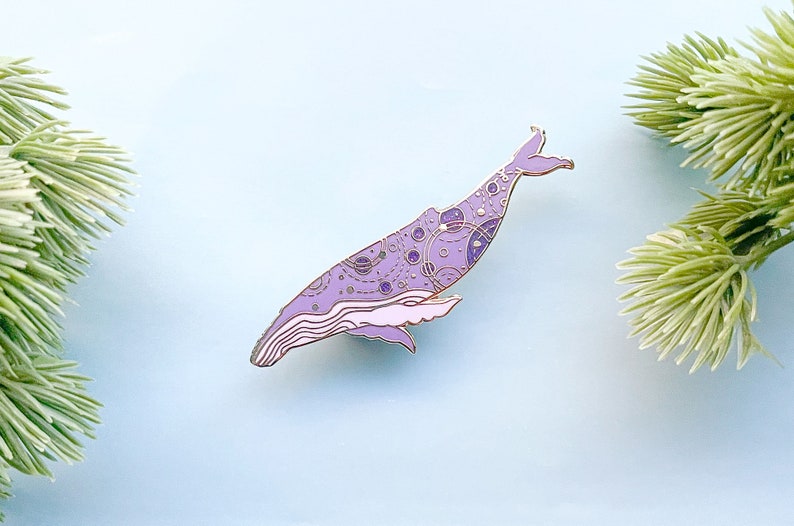 May include: A purple and gold enamel pin of a whale with a detailed pattern of circles and lines. The whale is in a swimming position with its tail raised.
