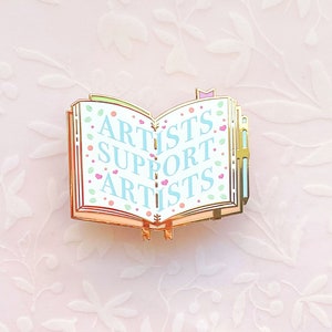 May include: Gold-tone enamel pin shaped like an open book with the words "Artists Support Artists" in blue and pink lettering on a white background.