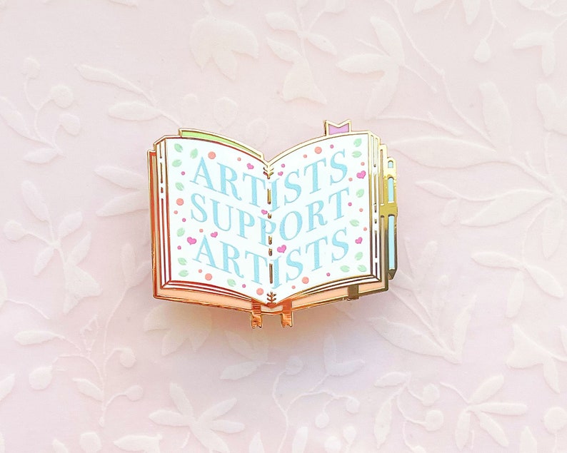 May include: Gold-tone enamel pin shaped like an open book with the words "Artists Support Artists" in blue and pink lettering on a white background.