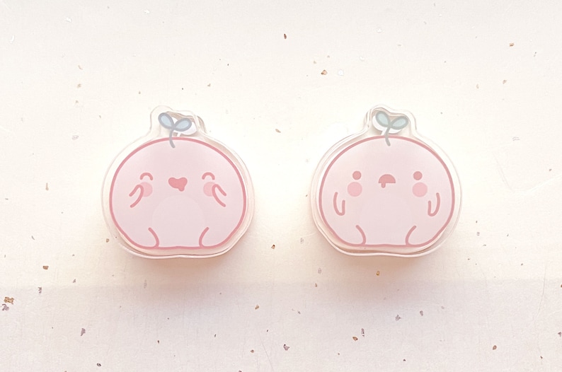 May include: Two pink cartoon character charms with a green leaf on top. One charm has a heart and a smile, the other has a frown and two dots for eyes.