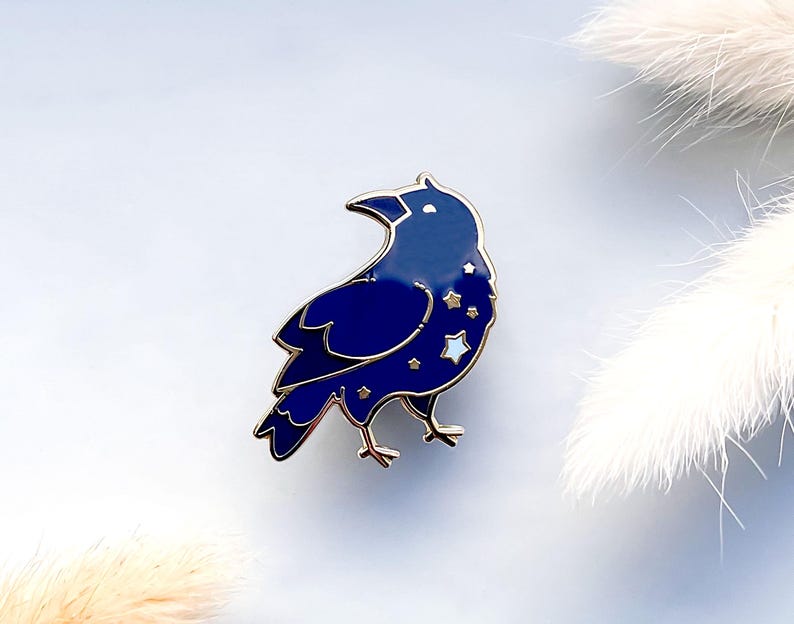 May include: A dark blue raven enamel pin with gold-colored accents. The raven is adorned with star-shaped details. The pin is a decorative accessory, suitable for jackets, bags, or other fabric items.