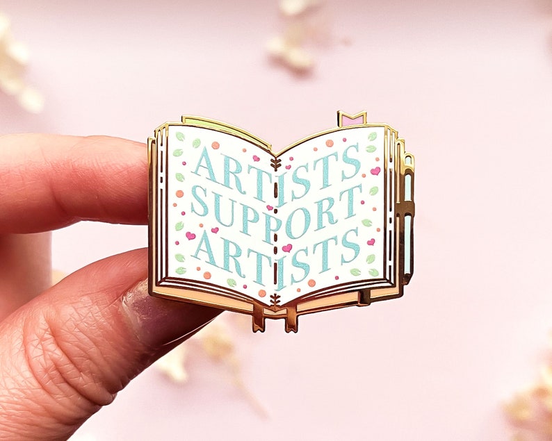 May include: Gold-toned enamel pin shaped like an open book with the words "Artists Support Artists" in blue text on a white background. The book has a floral pattern with pink hearts and green leaves.
