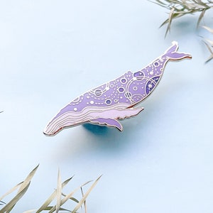 May include: A purple and white enamel pin of a whale with a detailed pattern of circles and lines. The whale is facing left and has a silver outline.