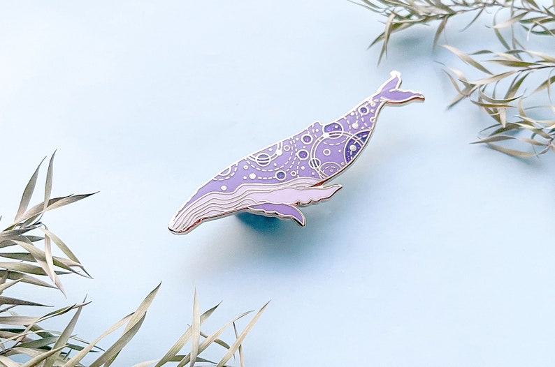 May include: A purple and white enamel pin of a whale with a detailed pattern of circles and lines. The whale is facing left and has a silver outline.