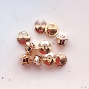 May include: A group of ten gold-toned metal pin backs. The pin backs are round with a small hole in the center. They are arranged in a pile on a white surface.