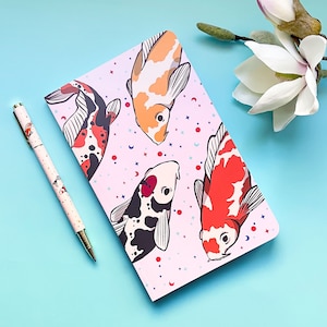 May include: A pink notebook featuring a colourful koi fish design. The notebook has a white background with a pattern of stars, moons, and dots. The koi fish are in various colours, including orange, red, black, and white.
