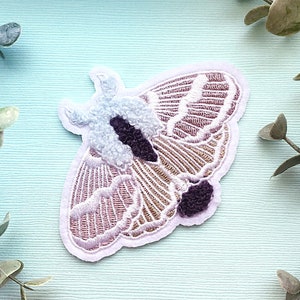 May include: A pink and white embroidered moth patch with a fluffy white body and purple accents. The patch is on a light blue background.
