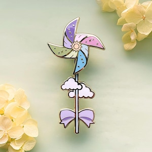 May include: A colorful pinwheel enamel pin with a gold-colored metal finish. The pinwheel features pastel colors, stars, and a sun in the center. Below the pinwheel are white clouds and a lavender bow. The pin is a whimsical accessory.