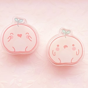 May include: Two peach-colored resin charms shaped like cartoon characters with small green leaves on top. The charms have simple facial features and are set against a pink background.