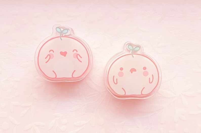 May include: Two peach-colored resin charms shaped like cartoon characters with small green leaves on top. The charms have simple facial features and are set against a pink background.
