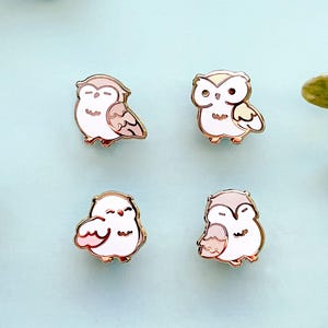 May include: Four enamel pins featuring cartoon owls in various poses. Each pin has a white body, with details in shades of pink, beige, and gray. The pins are outlined in gold and set against a light blue background.
