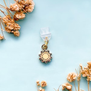 May include: A gold-toned metal charm shaped like a sun with a central circle and radiating points. The charm is attached to a small gold-toned jump ring, which is connected to a clear, translucent plastic plug-style attachment component.