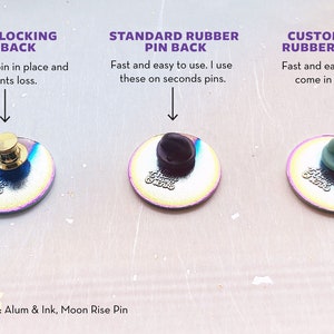 May include: Three different types of pin backs for enamel pins. The first is a metal locking pin back, the second is a standard rubber pin back, and the third is a custom color rubber pin back. The pin backs are shown on a rainbow iridescent enamel pin.