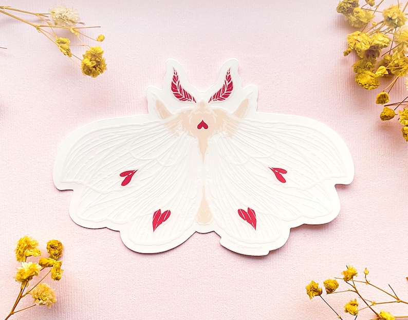 May include: White moth sticker with red hearts on its wings. The moth has a delicate, feminine design and is perfect for adding a touch of whimsy to any project.