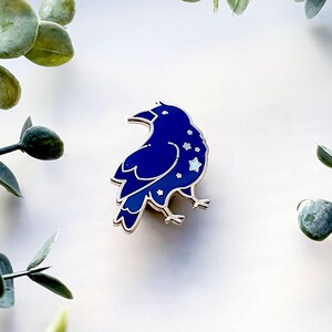 May include: A dark blue enamel pin in the shape of a raven, with a silver outline. The raven has a starry design on its body. The pin is set against a white background with green foliage.