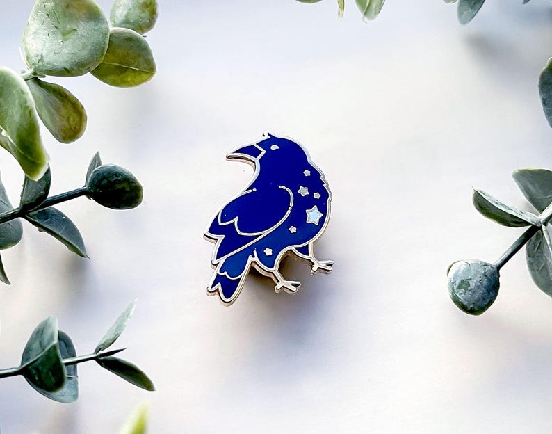 May include: A dark blue enamel pin in the shape of a raven, with a silver outline. The raven has a starry design on its body. The pin is set against a white background with green foliage.