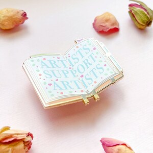 May include: Gold-toned enamel pin shaped like an open book with the words "Artists Support Artists" in blue and pink lettering on a white background with pink hearts.