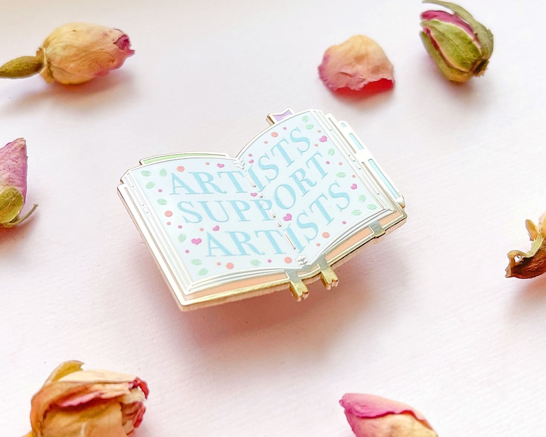 May include: Gold-toned enamel pin shaped like an open book with the words "Artists Support Artists" in blue and pink lettering on a white background with pink hearts.