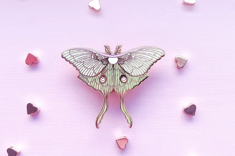 Puede incluir: Un pin de metal verde y dorado de una polilla luna con alas detalladas y un cuerpo rosa. El pin est&aacute; sobre un fondo rosa con peque&ntilde;os corazones dorados.