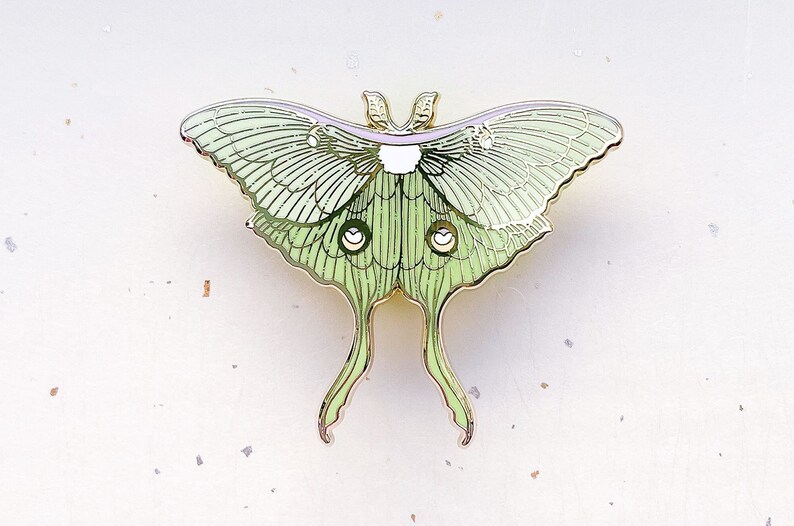 Puede incluir: Un pin de metal dorado con una polilla luna verde y blanca con alas y antenas detalladas.